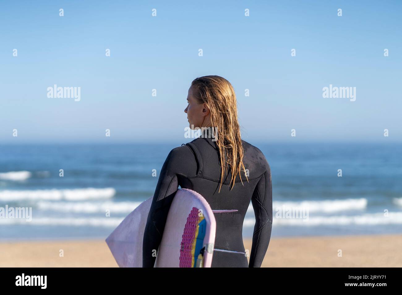 Surfer girl at the beach with her surfboard looking at the ocean waves in the morning. Stock Photo