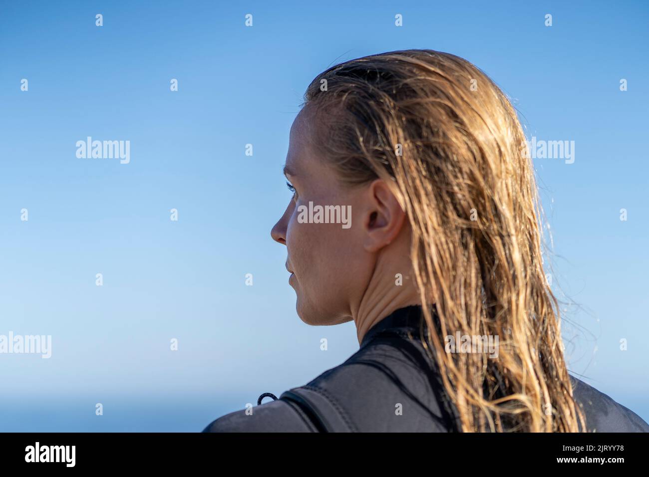 Surfer girl with wet hair in the morning light at the beach looking at the waves. Stock Photo