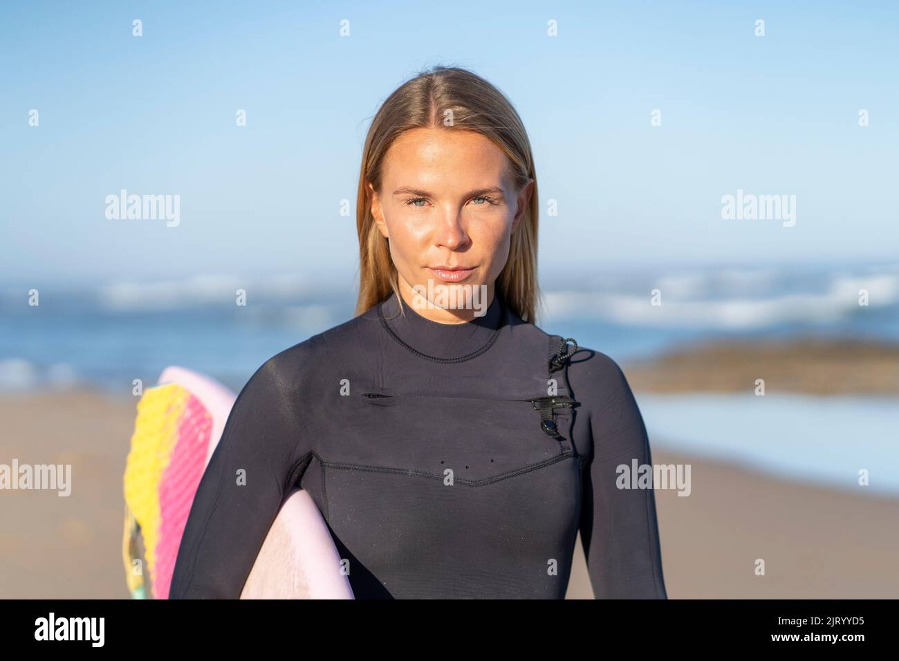 Surfer girl at the beach with her surfboard looking at the camera in the morning. Female surfer woman Stock Photo