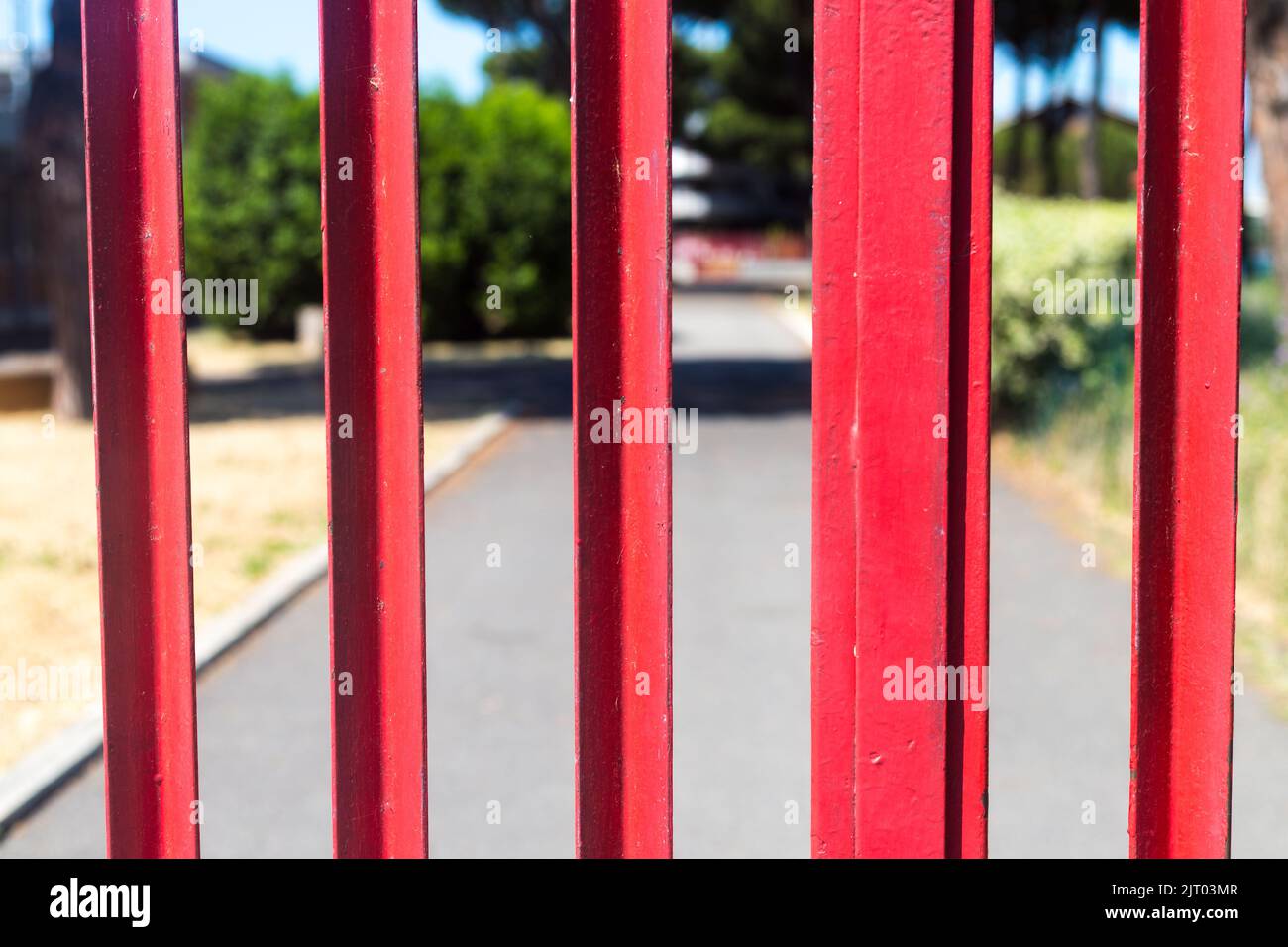 Closeup of closed red gate. School park closed for visitors. Asphalt road and alley behind a metal hence. Summer sunny day. School closed till Stock Photo