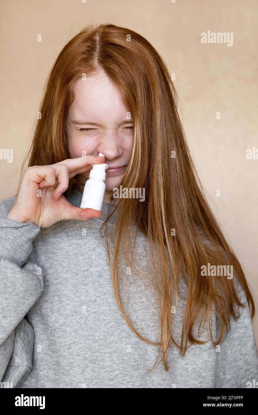 Beautiful European funny teenage girl with long red hair in a grey hoodie squinting her nose with a cure for the common cold from a white bottle Stock Photo