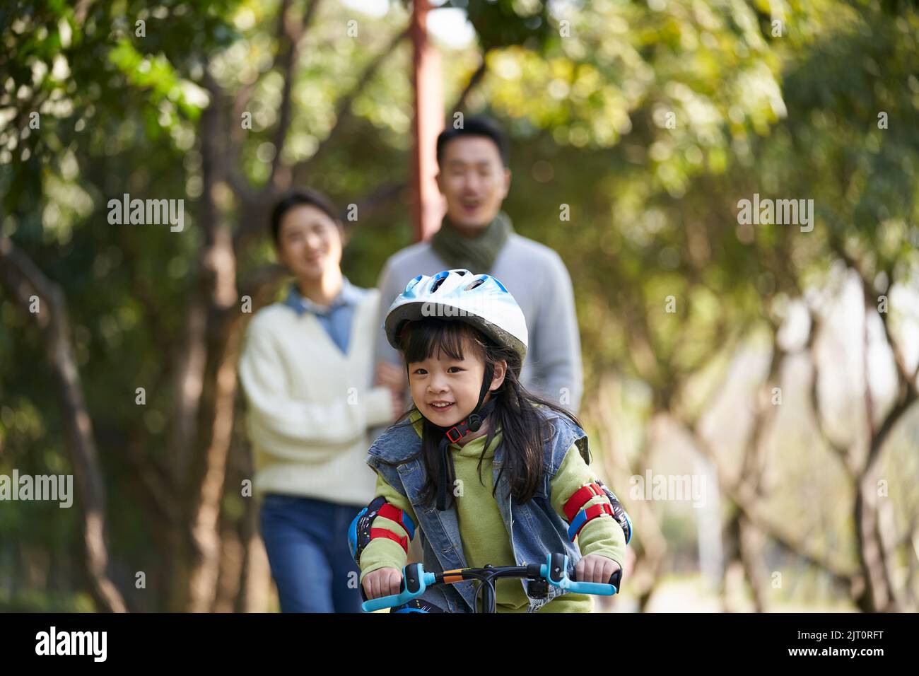little asian girl riding bike in city park with parents in background Stock Photo