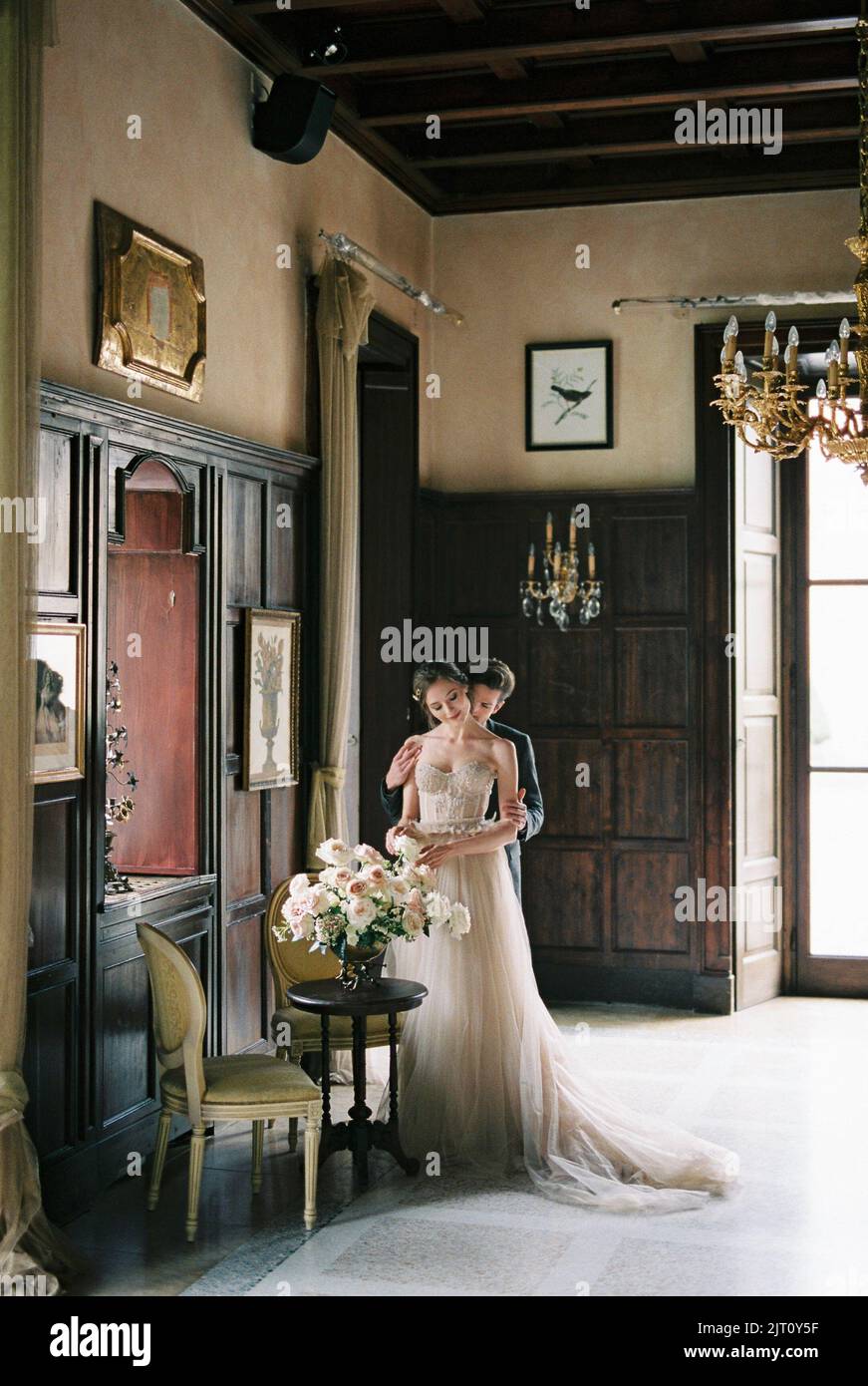 Groom hugs bride from behind in a room near the window of an old villa ...