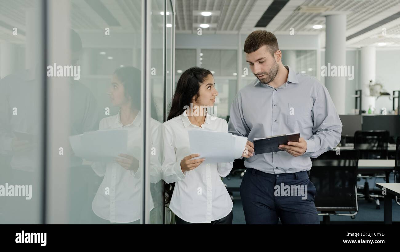 Business young man and woman colleagues stand in office communicate discuss joint project girl trainee asks clarifies details task talks with mentor Stock Photo
