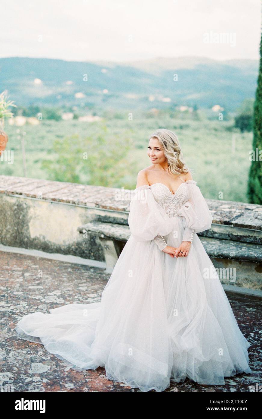 Bride in a white puffy dress stands near a stone fence in the garden ...