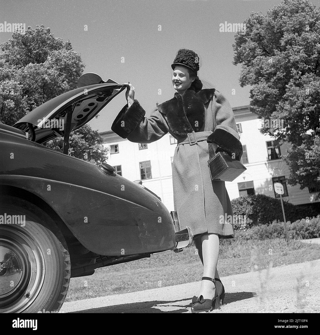 Women's fashion in the 1940s. A young woman in a typical 1940s outfit. A warm coat, matching shoes, handbag and hat. She poses the outfit beside a car and an open trunk. Sweden 1946. Kristoffersson Ref U129-3 Stock Photo
