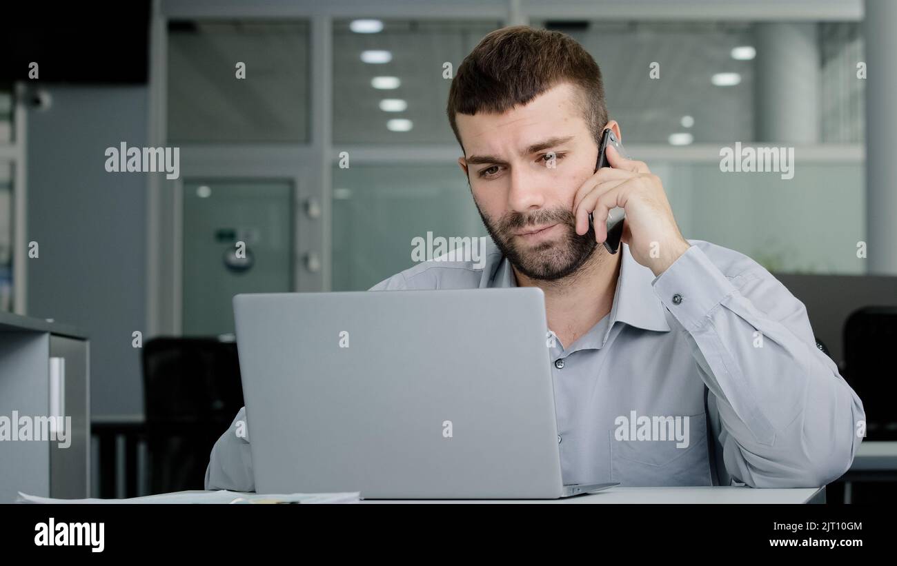 Concentrated young businessman speaks on phone consults client checks data on laptop screen serious manager remote seller offers services over Stock Photo