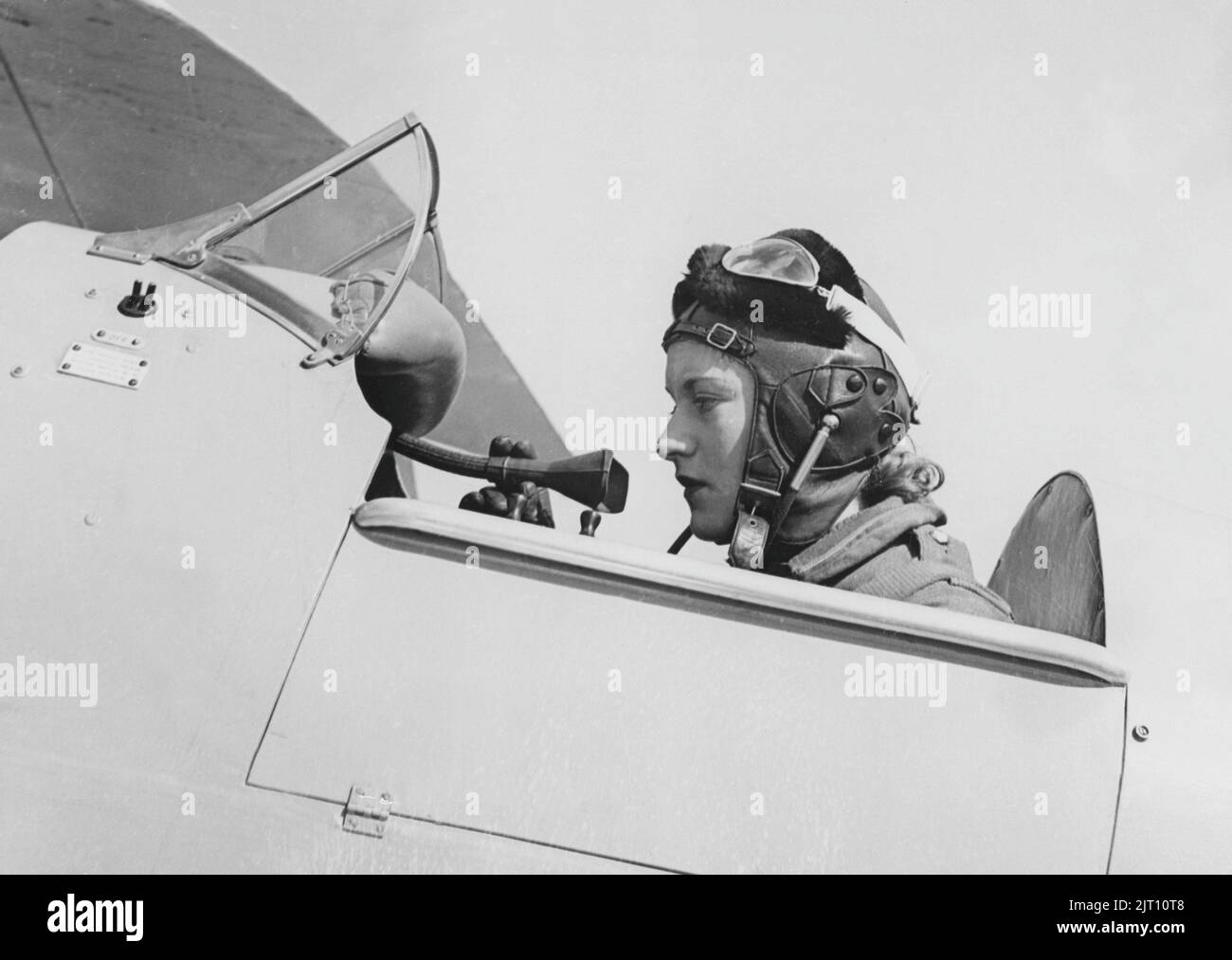 In the 1930s. A young female pilot is seen sitting in the cockpit of her airplane wearing the leather cap and glasses ready for takeoff. 19399 Stock Photo