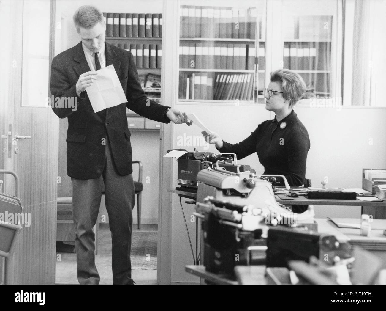 Office girl in the 1960s. A woman is sitting by her typewriter and a man is standing in the room both reading and being handed a document from her at the same time. A good illustration of how the relations were at the time between employees and managers as he gives her not attention or gratitude for her work, visibly a man who consider himself being on a higher level in the company than her. Sweden 1960. Stock Photo