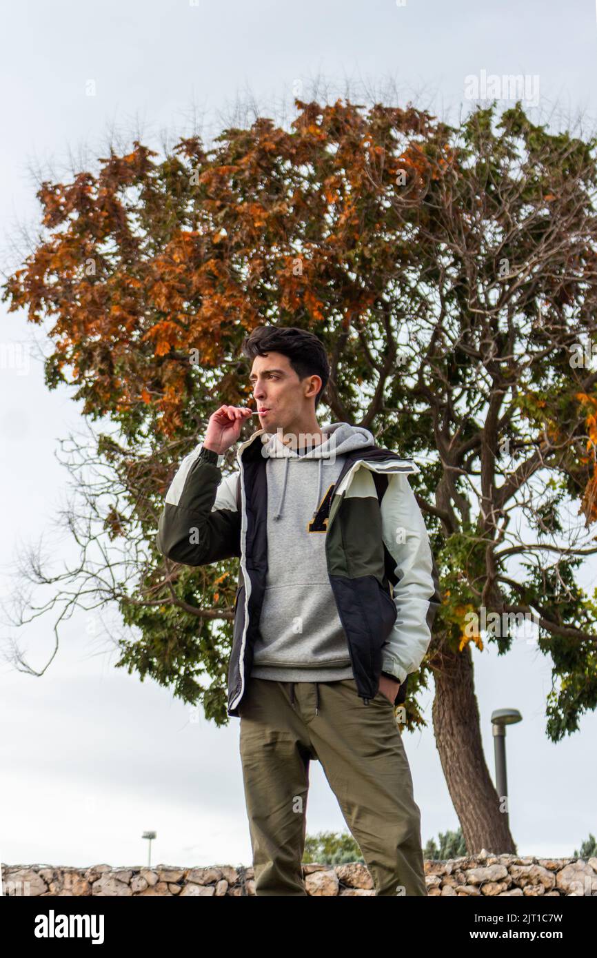 A handsome caucasian man eating a red lollipop posing in a park with casual attire. Its autumn Stock Photo
