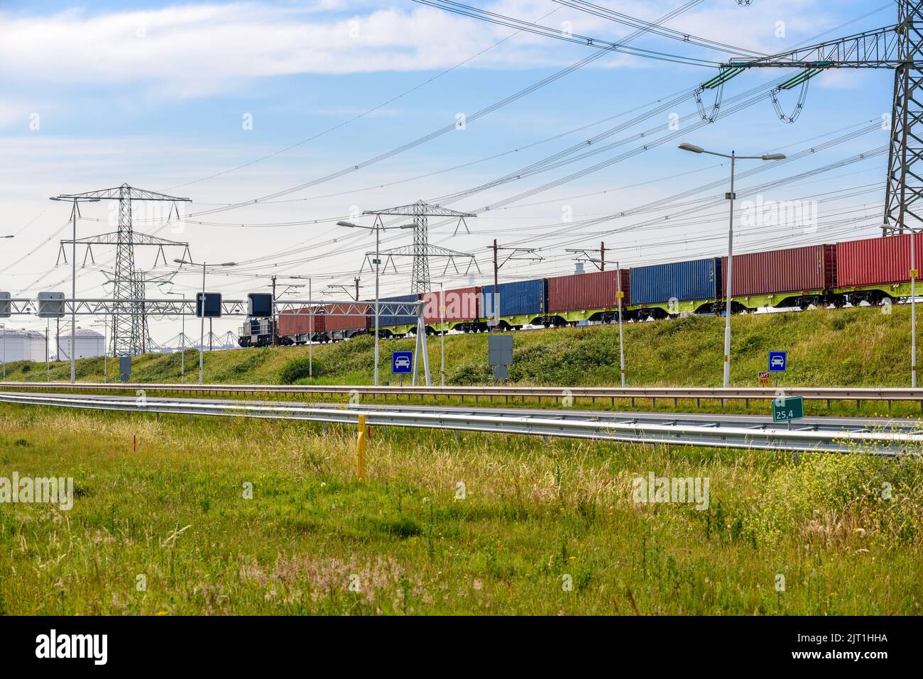 Cargo train loaded with containers running along a motorway in a port area on a clear summer day. Electricity pylons and fuel tanks are in background. Stock Photo