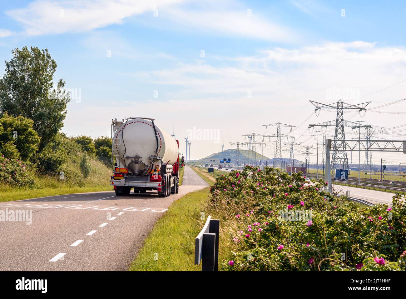Bulk tanker lorry on a narrow road running along a motorway and railway in a port area on a sunny summer day. Electricity pylons are in background. Stock Photo