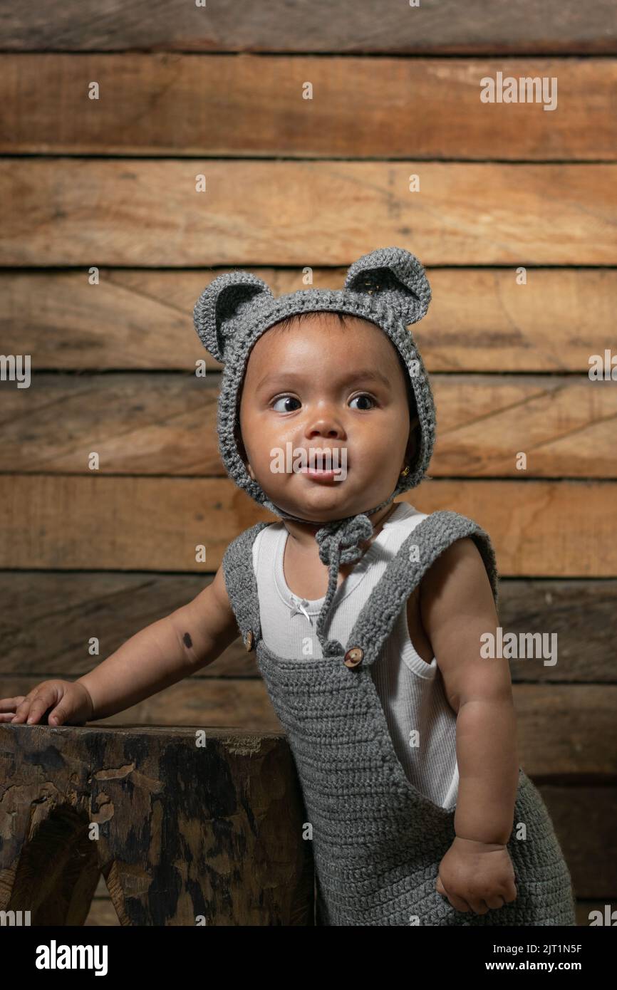 beautiful baby latina with brown skin, standing on a wooden chair and an orange background, learning to take her first steps, wearing overalls and a c Stock Photo