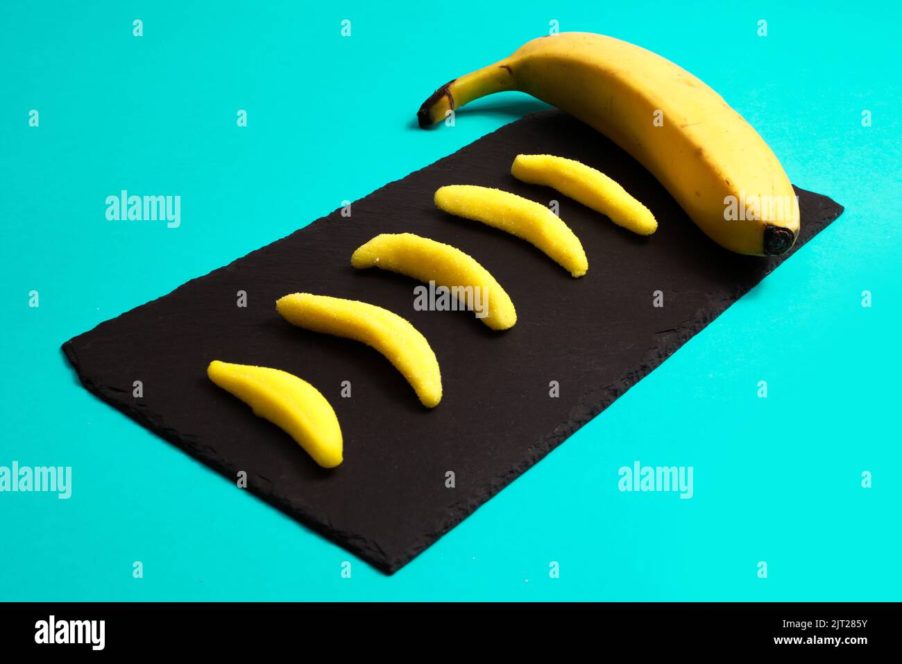 Small banana-shaped candies and a real banana on a black tray isolated on turquoise background Stock Photo