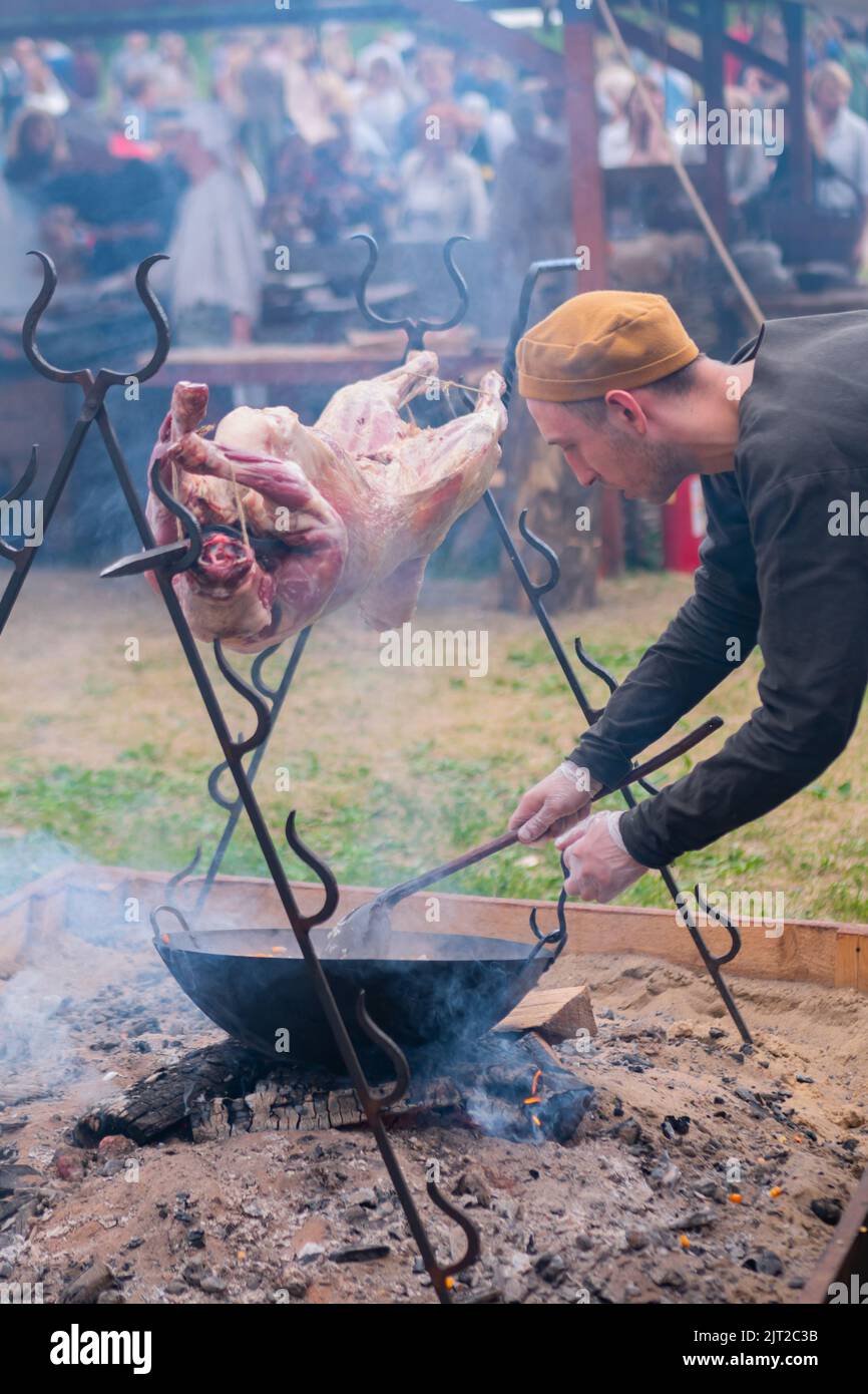 Man cooking roasted pork carcass on spit and vegetables in pot over open fire Stock Photo