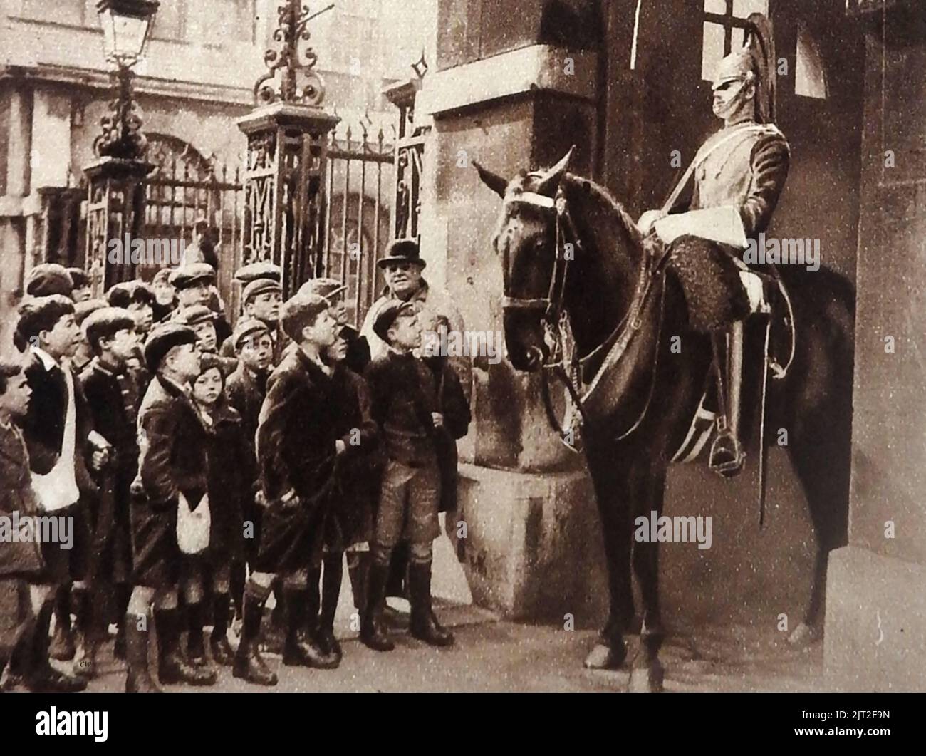 OLD LONDON - A vintage photo showing a group of visiting schoolchildren meeting a horse guard at Horse Guards Parade. All the boys are wearing short trousers and flat caps, once an often  traditional fashion in Britain , worn by boys under the age of eleven before they attended senior school. Stock Photo