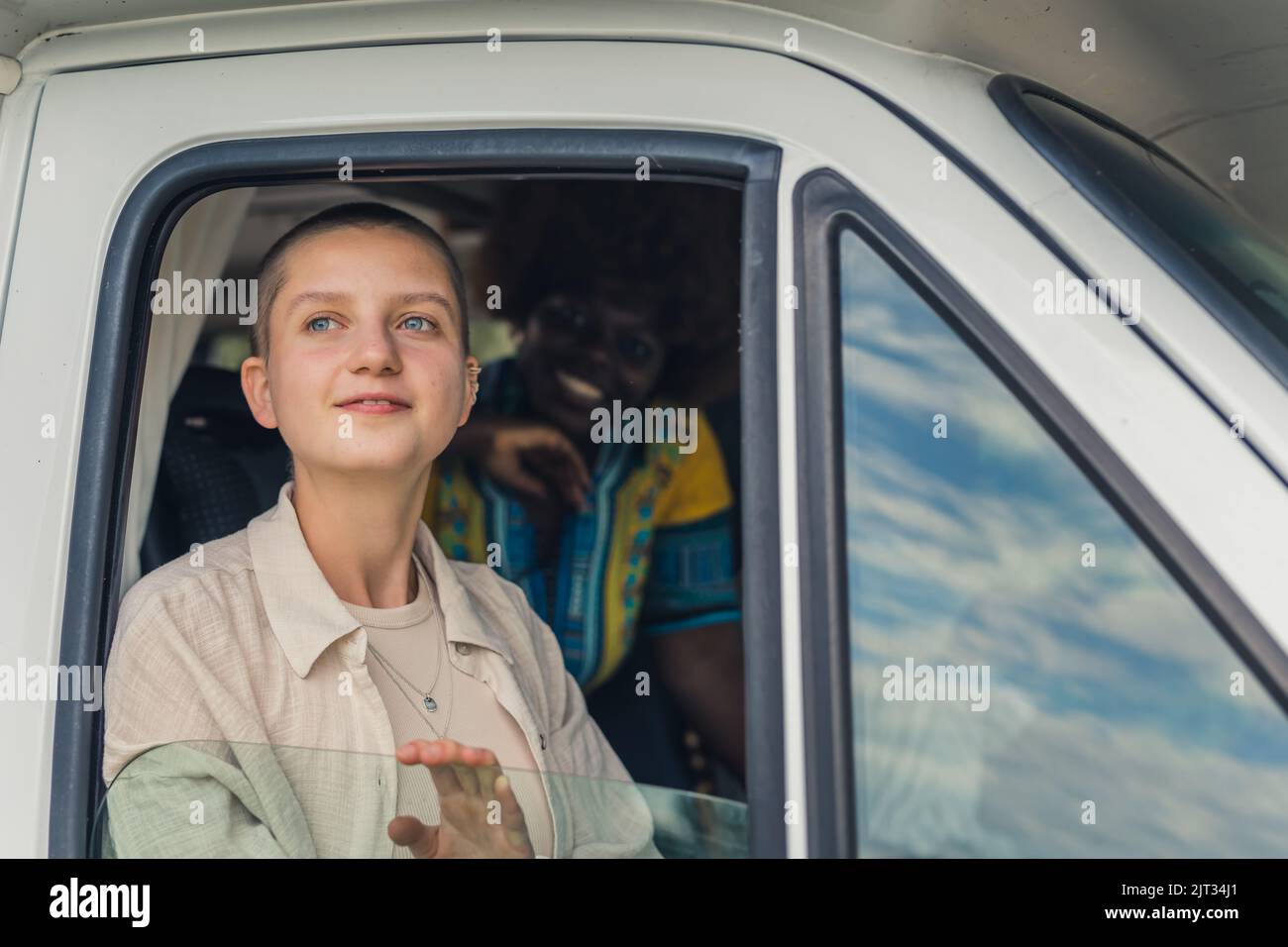 Young caucasian beautiful girl with shaved head looking out of the open window of a camping van in awe. Her smiling African young woman friend with kinky hair on the back seat. High quality photo Stock Photo