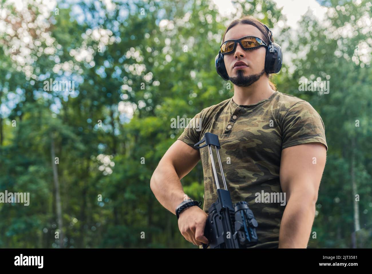 Medium outdoor shot of a military-looking caucasian muscular man in moro t-shirt looking far away at his target and holding black rifle with a loupe to look through. High quality photo Stock Photo