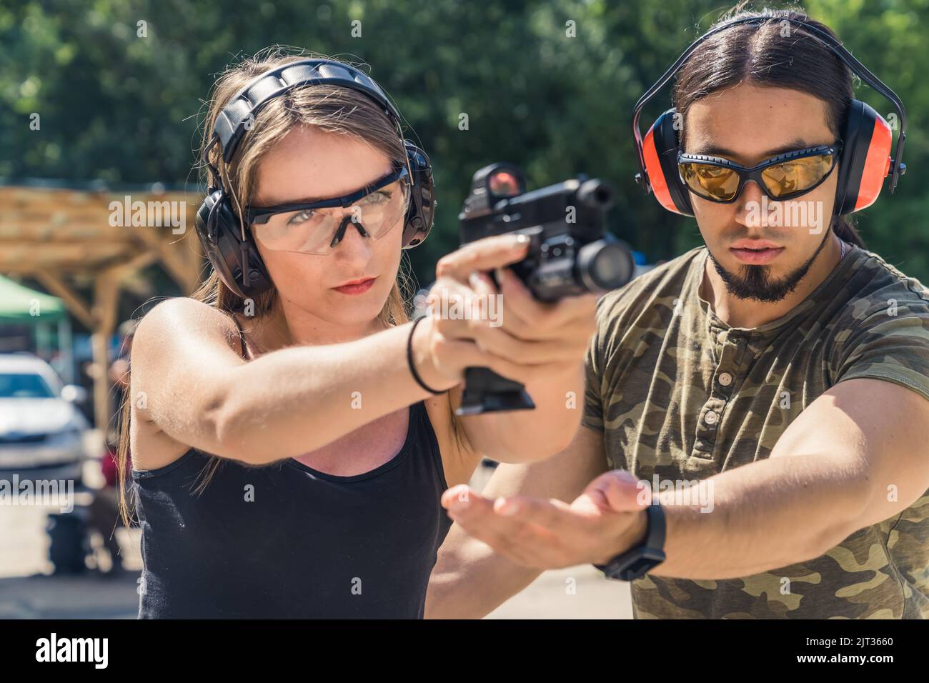 Outdoor gun range concept. Two caucasian adults - a man and a woman. Male instructor in protective gear in moro t-shirt helping out with his client's posture. High quality photo Stock Photo