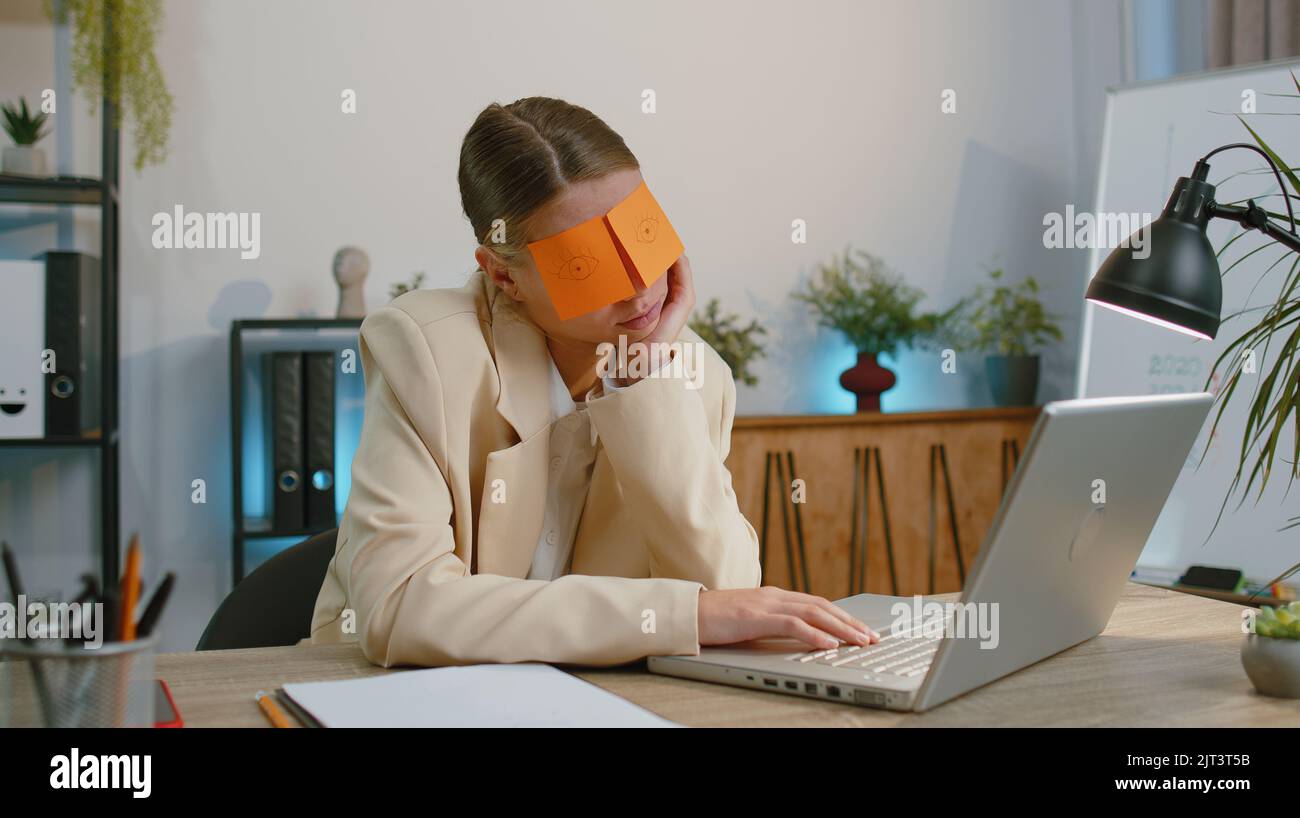 Inefficient tired businesswoman in suit working sleeping on laptop computer with eyes stickers on face at office workplace desk. Funny lazy manager freelancer woman. Business people. Cheating to sleep Stock Photo