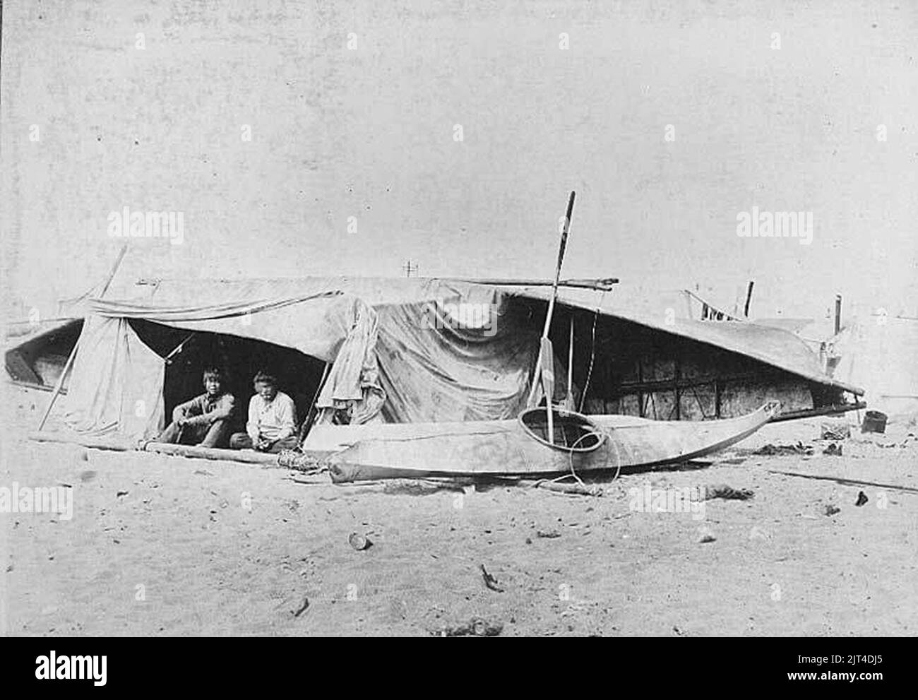 Two Inuit men sitting beneath a umiak turned on its side, next to a