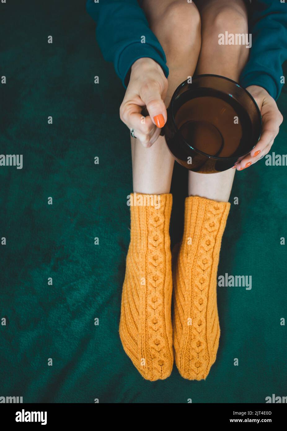 Girl in orange knitted socks with cup of tea, top view. Cozy time at home. Winter time. Woman sitting on green sofa with hot tea, top view. Stock Photo