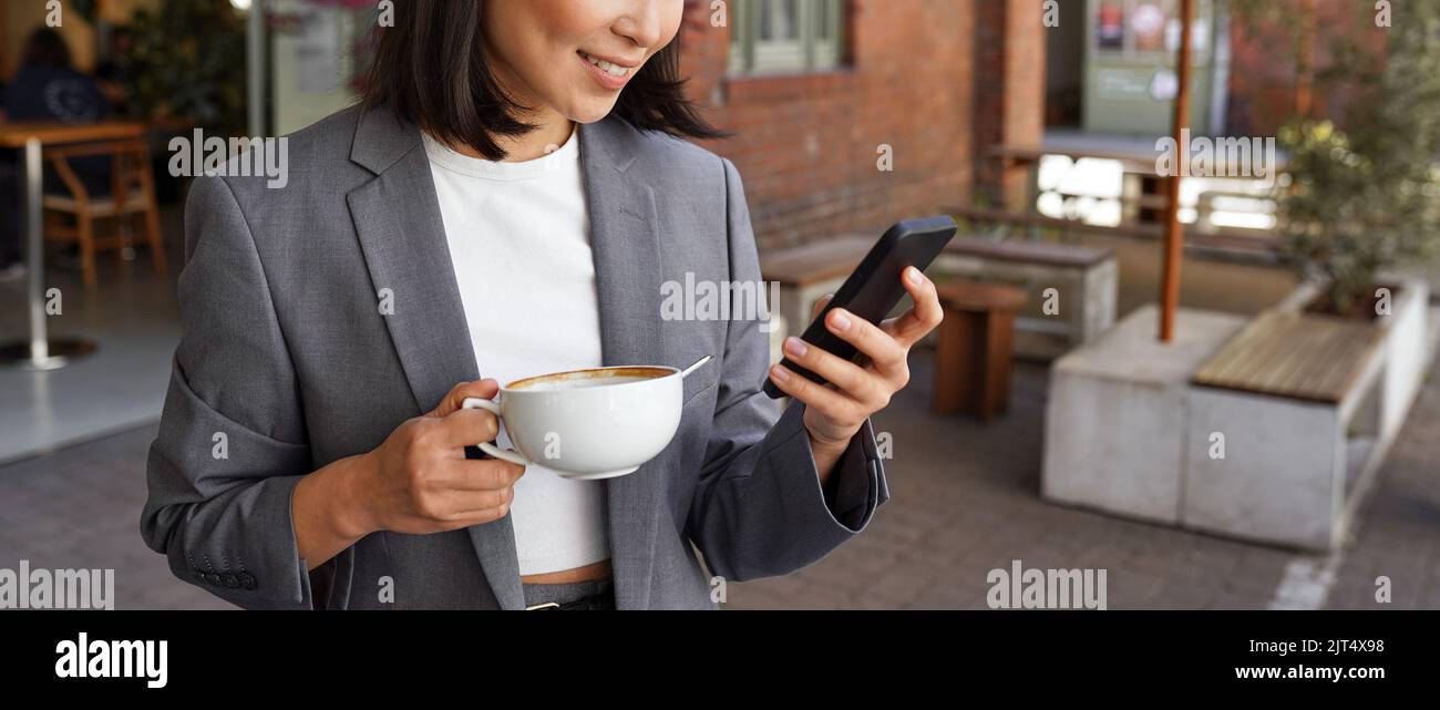 Young elegant professional woman holding coffee standing outside using phone. Stock Photo