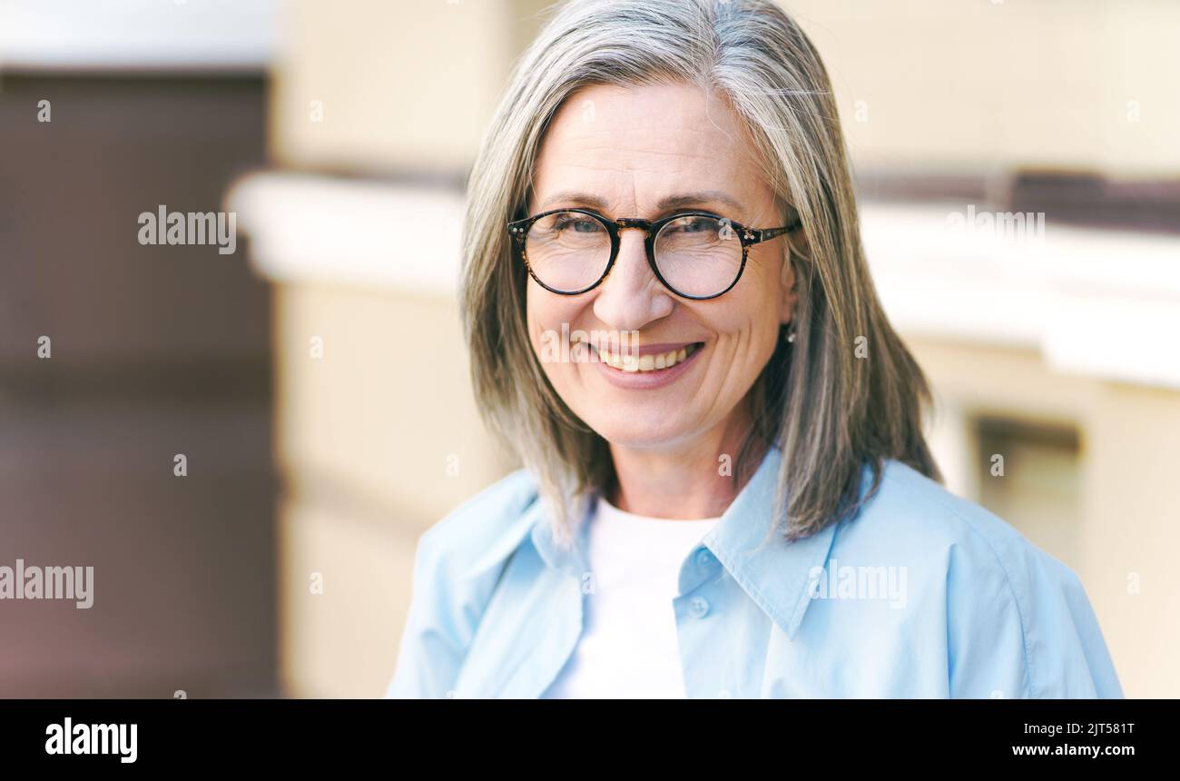 Charming grey hair mature woman wearing eye glasses standing outdoors enjoying vacation traveling around world at retirement. Mature woman with perfect skin wearing blue shirt.  Stock Photo