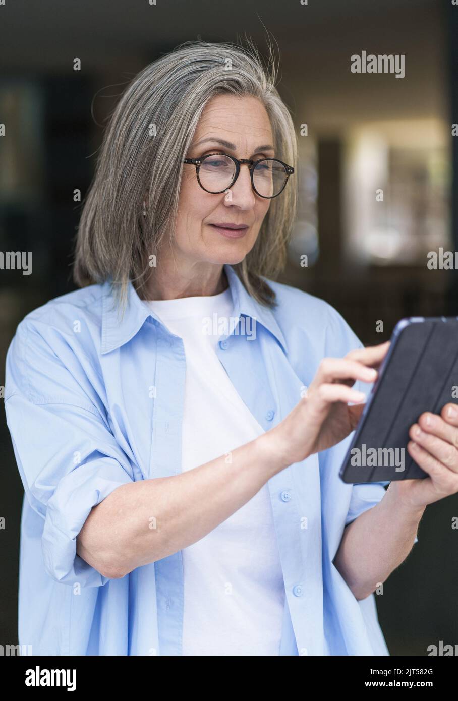 Interested in reading news or social media mature grey hair woman holding digital tablet sitting standing outdoors at the city streets. Mature woman answering call outdoors.  Stock Photo