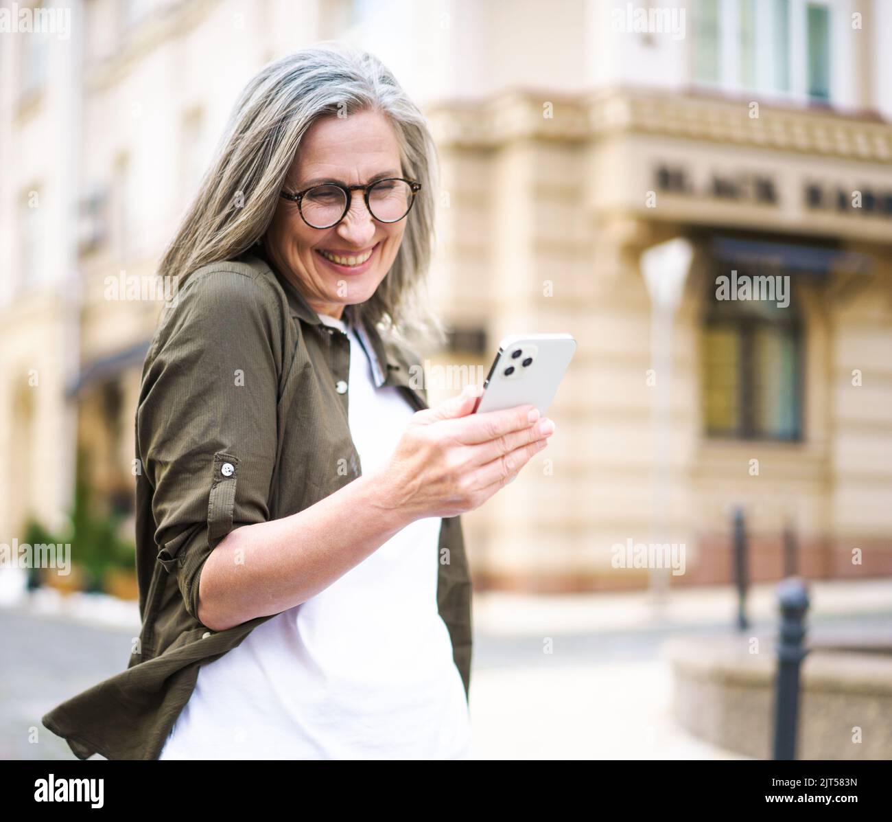 Mature beautiful grey hair woman looking at smartphone walking at the streets of old European town. Senior businesswoman read text message, answering video call standing outdoors. Selective focus. Stock Photo