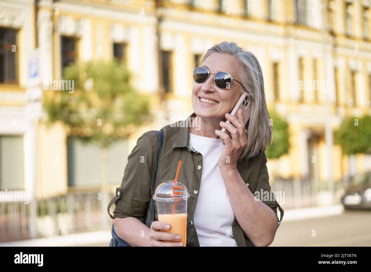 Answering phone call happy mature woman talking on the phone enjoying free time after work or traveling having juice in plastic cup on the go in city background. Enjoying life mature woman.  Stock Photo