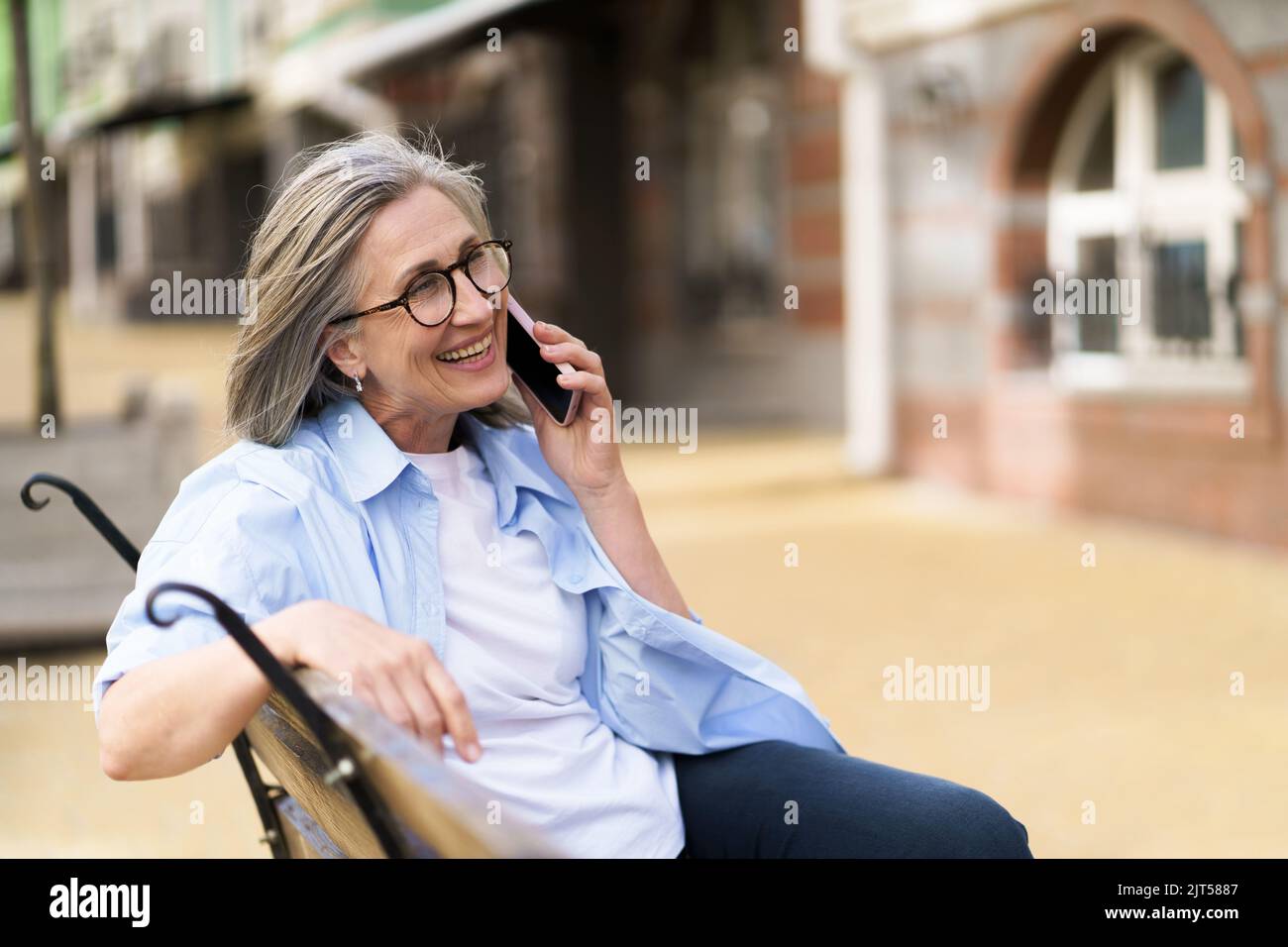 Mature grey hair woman smiling talking on the phone sitting on the bench enjoying summer time at the streets of old European town. Mature woman answering call outdoors.  Stock Photo