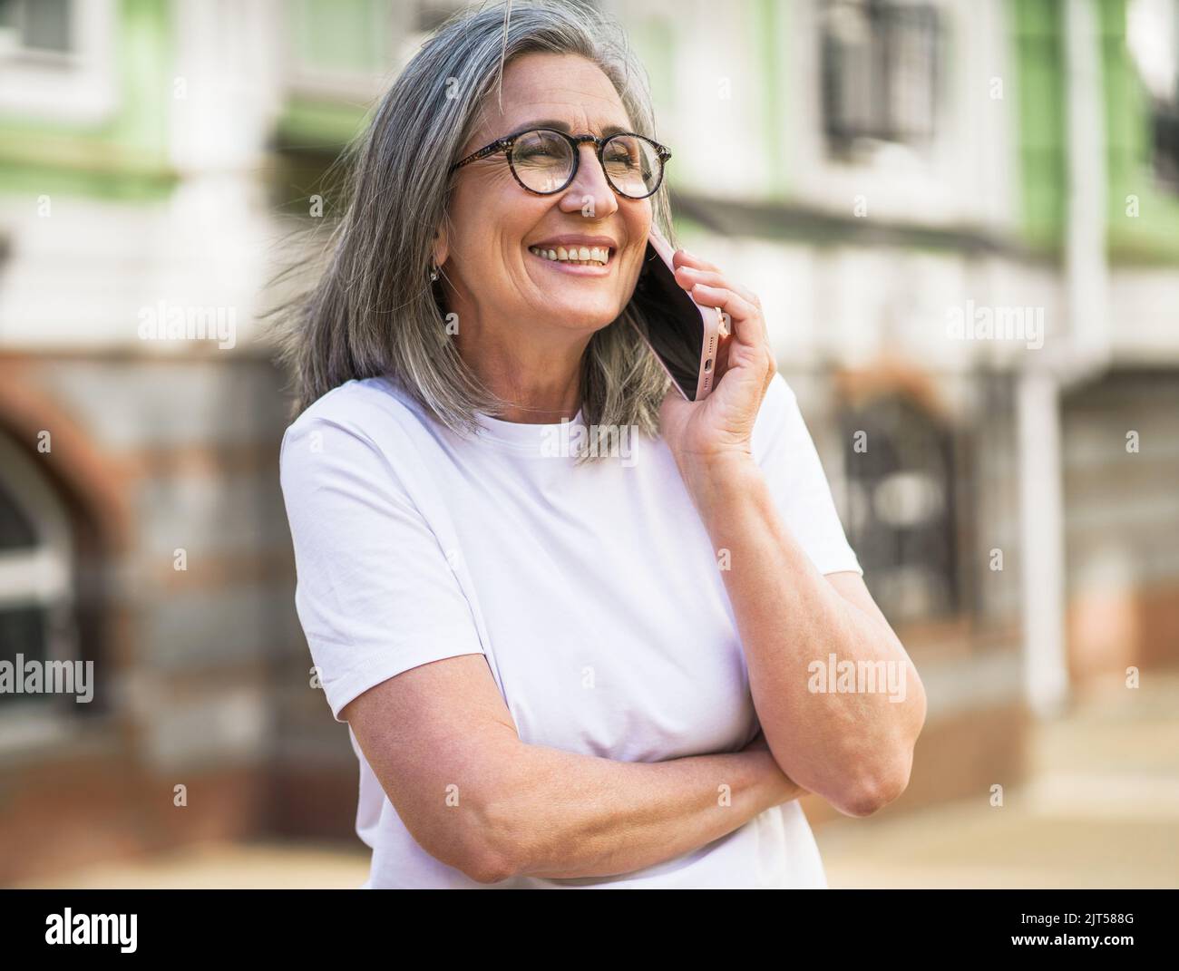 Mature grey hair business woman in eye glasses talking on the phone standing outdoors of the streets of old urban city wearing white t-shirt. Silver hair woman outdoors.  Stock Photo