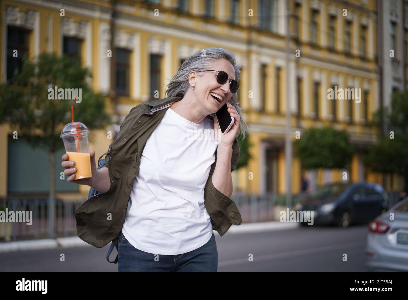 Happy mature European woman laughing talking on the phone enjoying free time after work or traveling having juice on the go using plastic cup in city background. Enjoying life mature woman.  Stock Photo