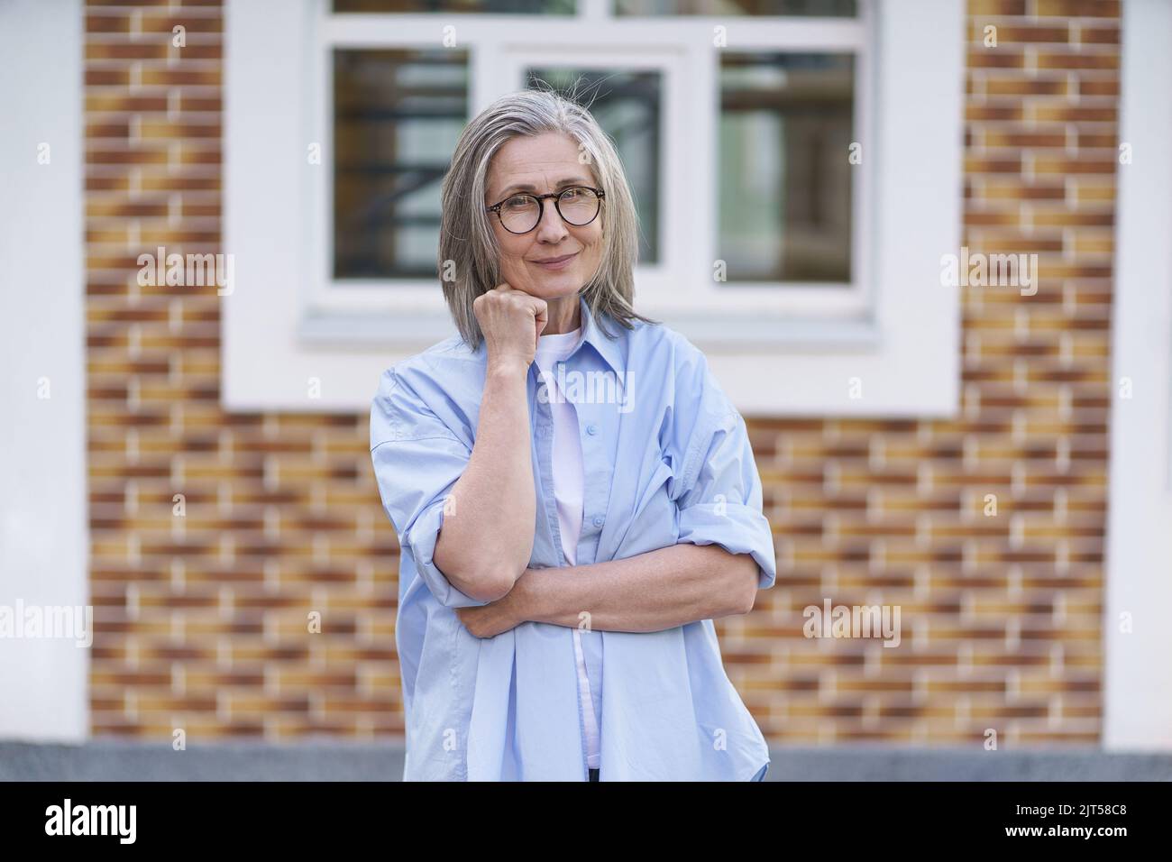 Mature grey hair woman wearing glasses standing outdoor traveling around world at retirement. Senior woman meeting her family. Mature woman with perfect skin wearing blue shirt standing outside.  Stock Photo