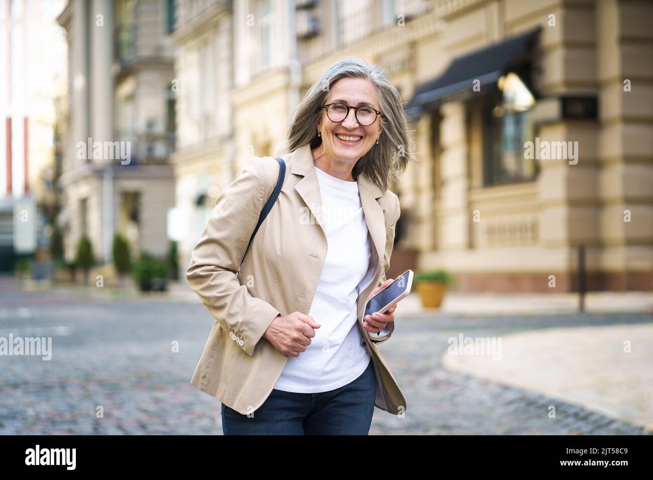 Mature elegant beautiful businesswoman. Senior woman in casual formal wear hold phone in hand walking on street from work or back to office. Mature woman spend free time traveling in European city.  Stock Photo
