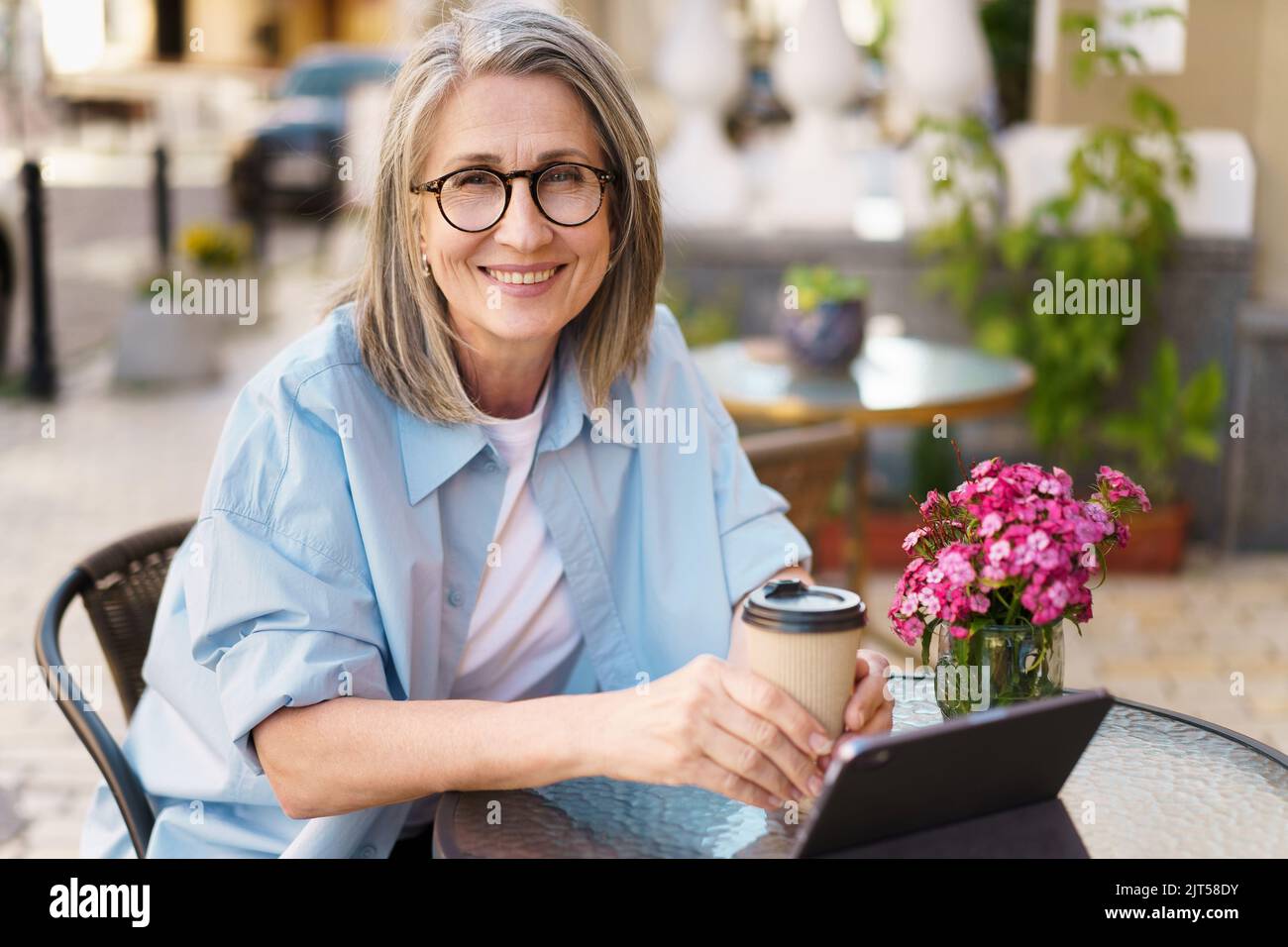 Mature European grey hair charming woman drink coffee using digital tablet sitting at table of street cafe. Mature silver hair woman working outdoors city cafe, wearing white t-shirt and blue shirt.  Stock Photo
