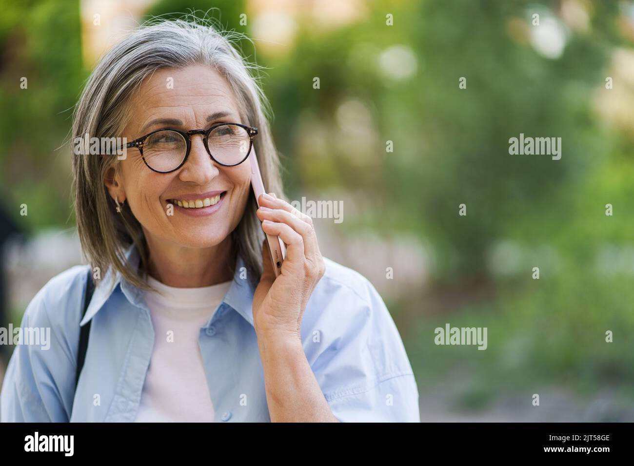 Senior grey hair woman in eye glasses smiling talking on the phone standing outdoors garden or park background wearing blue shirt. Silver hair woman outdoors.  Stock Photo