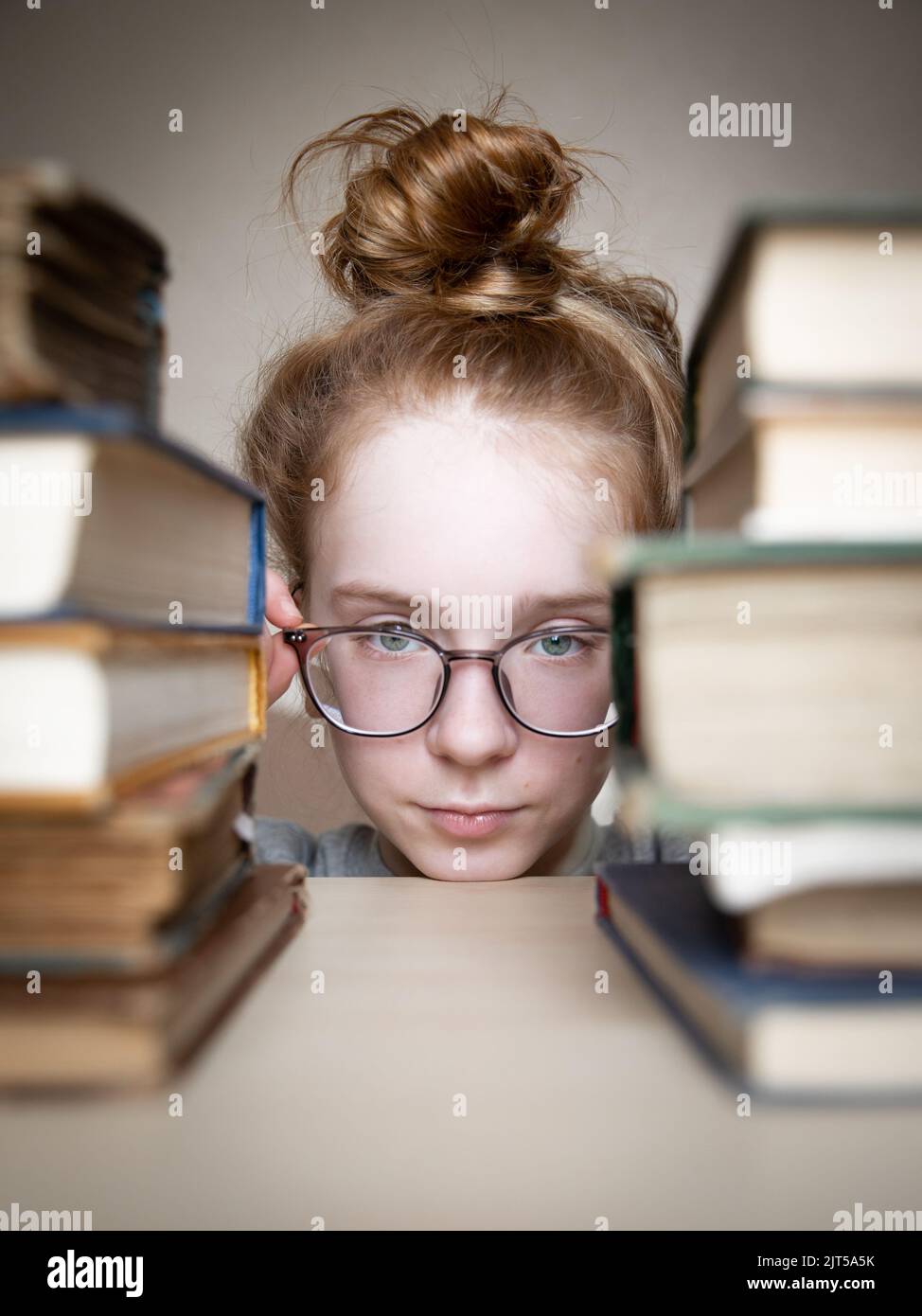 Beautiful teenage girl with red hair tied in bun looks between two stacks of hardback books lying on light table. Girl holding glasses on her face Stock Photo