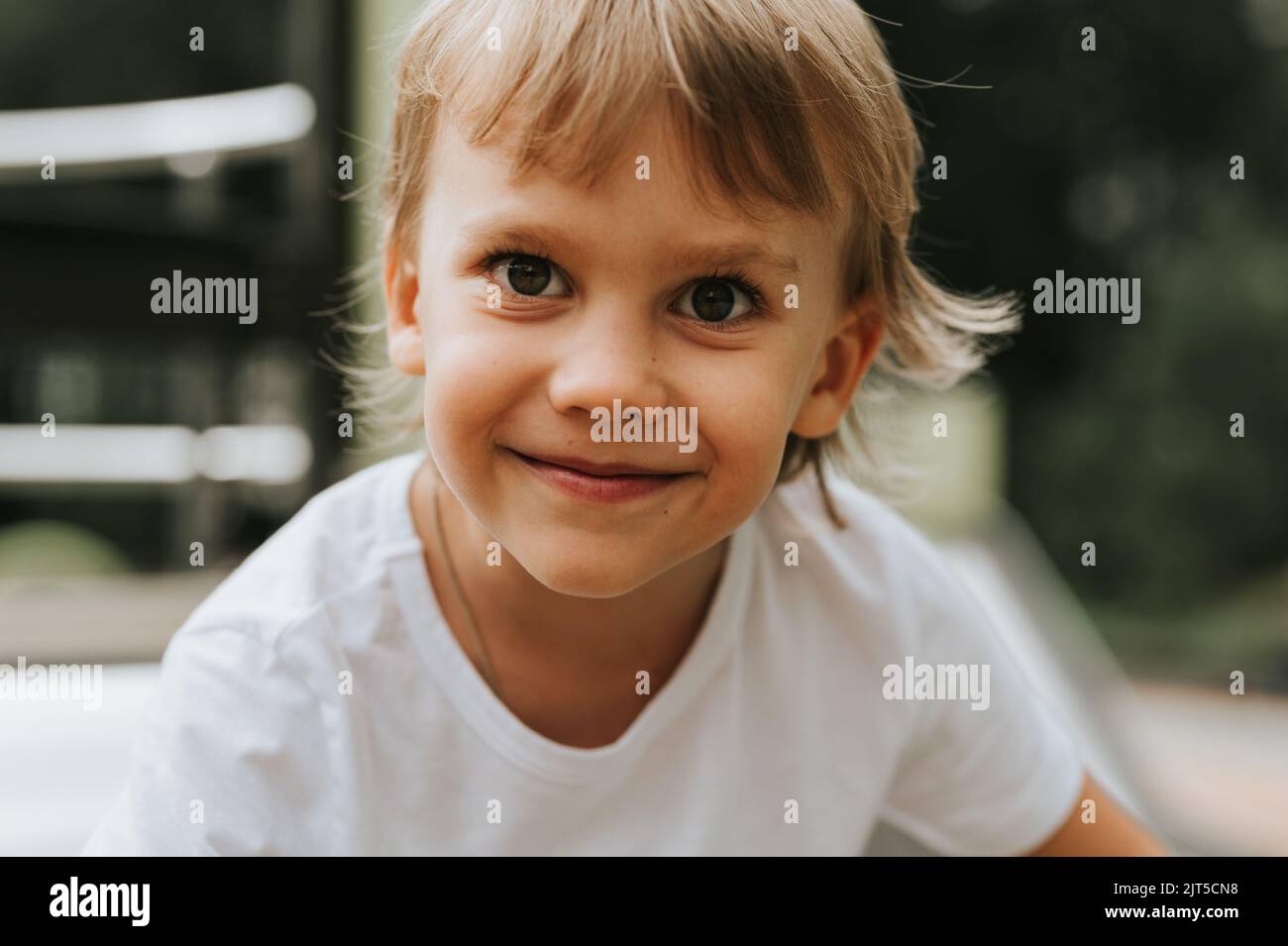 portrait of the face of a cute little happy smiling candid five year old kid boy with big eyes and long blond hair in a white t-shirt. generation z ch Stock Photo