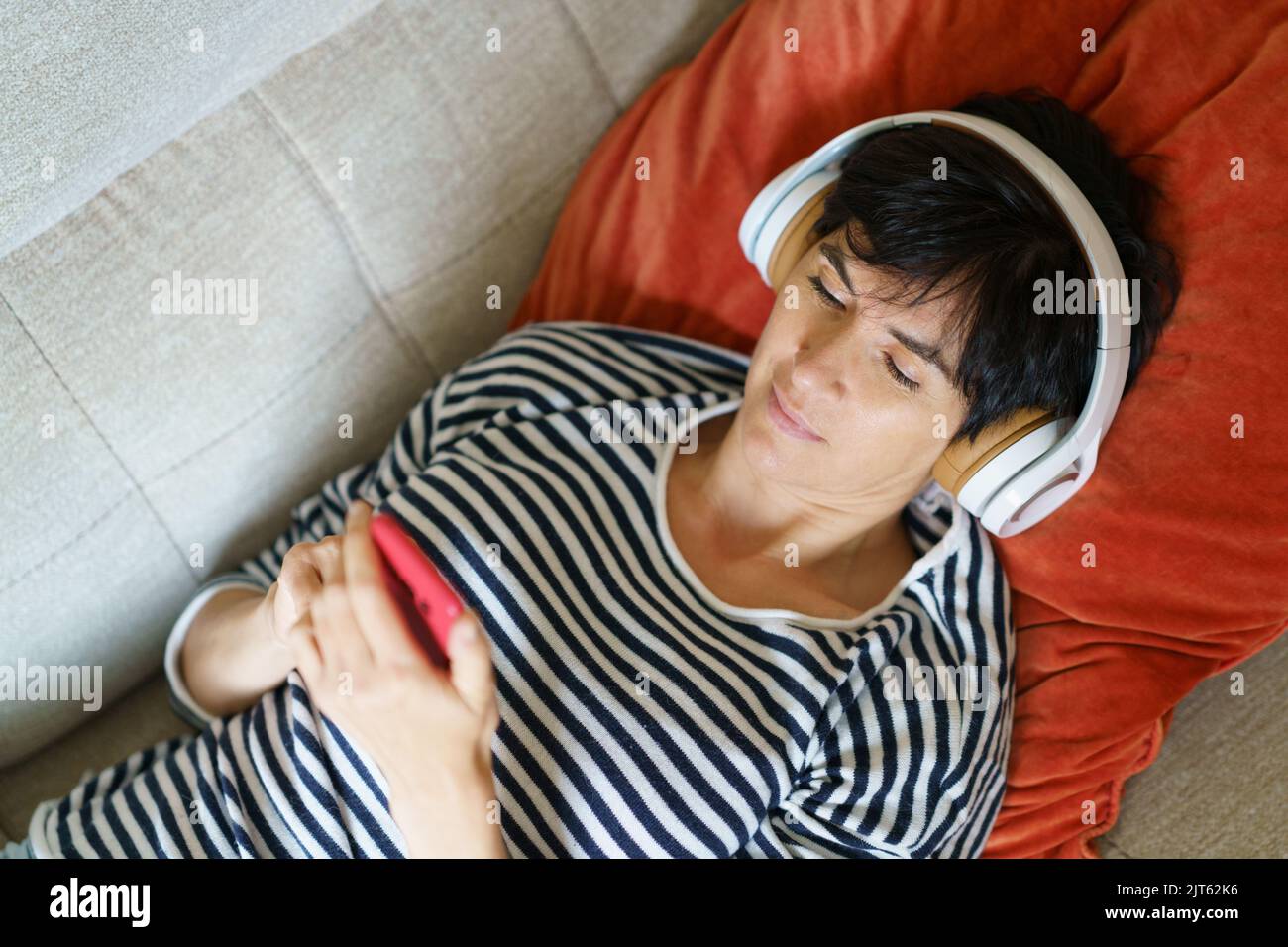 Woman consulting her smartphone while listening to music with headphones lying on the sofa. Stock Photo