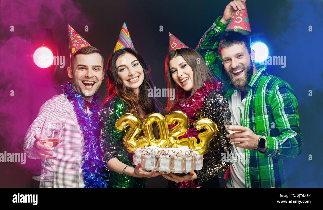 A company of young adult friends in party hats hold a New Year's birthday cake with numbers 2023 in their hands. Stock Photo