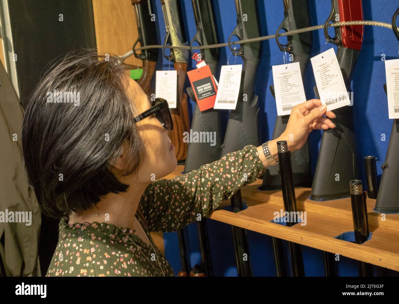 An Asian woman customer looks at 12-bore shotguns for sale in the shop at Southdown Gun Club & Shooting School, Findon, West Sussex, UK. Stock Photo