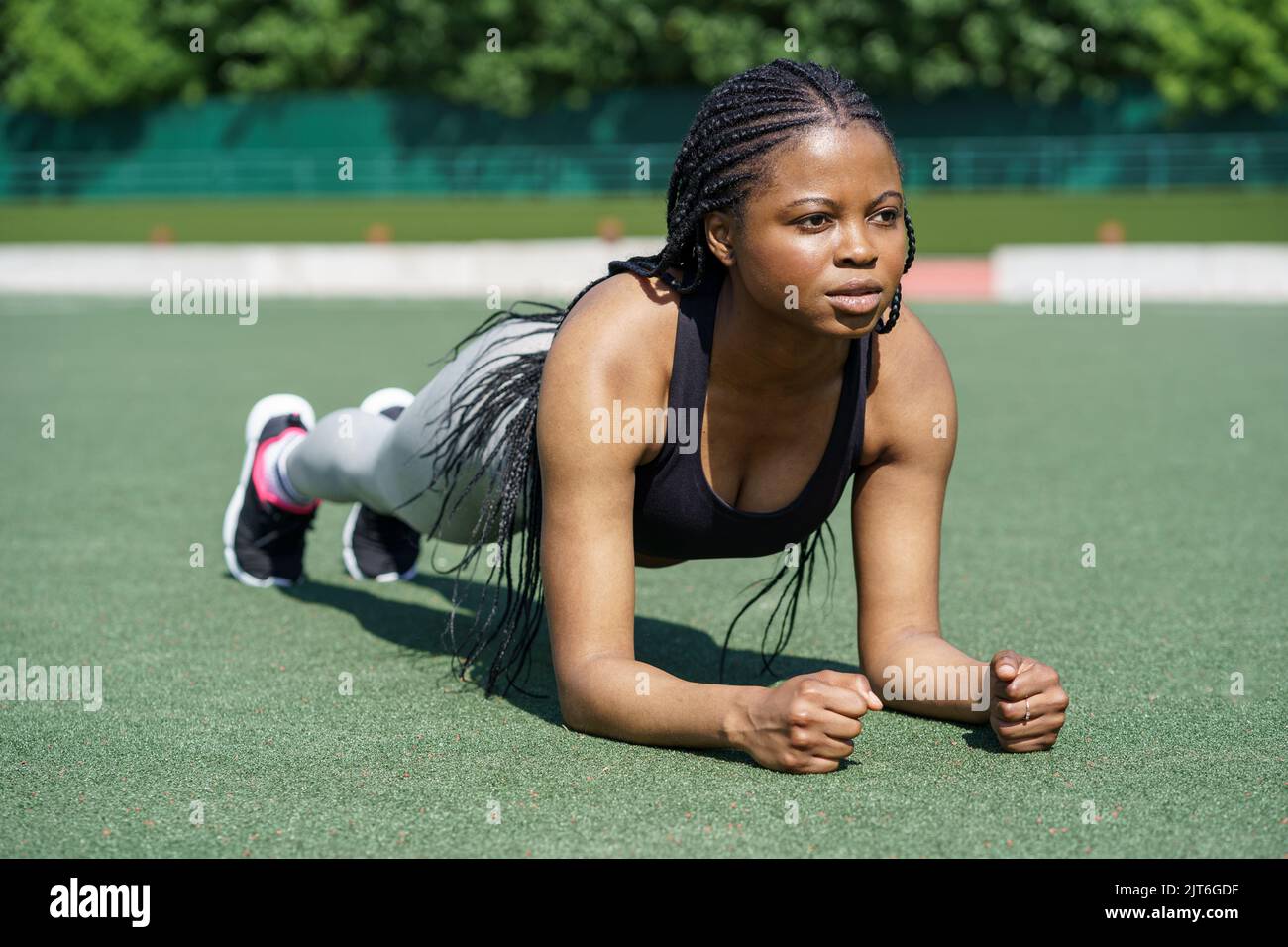 Determinated black woman does plank exercise on green turf flooring of city stadium against park Stock Photo