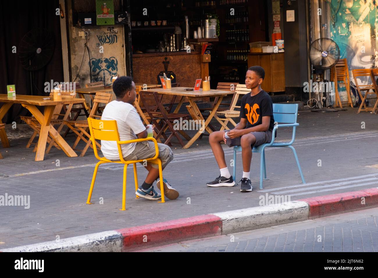 Black boys sitting on contrasting chairs, exchanging glances on Jaffa street, with bar and restaurant backdrop Stock Photo