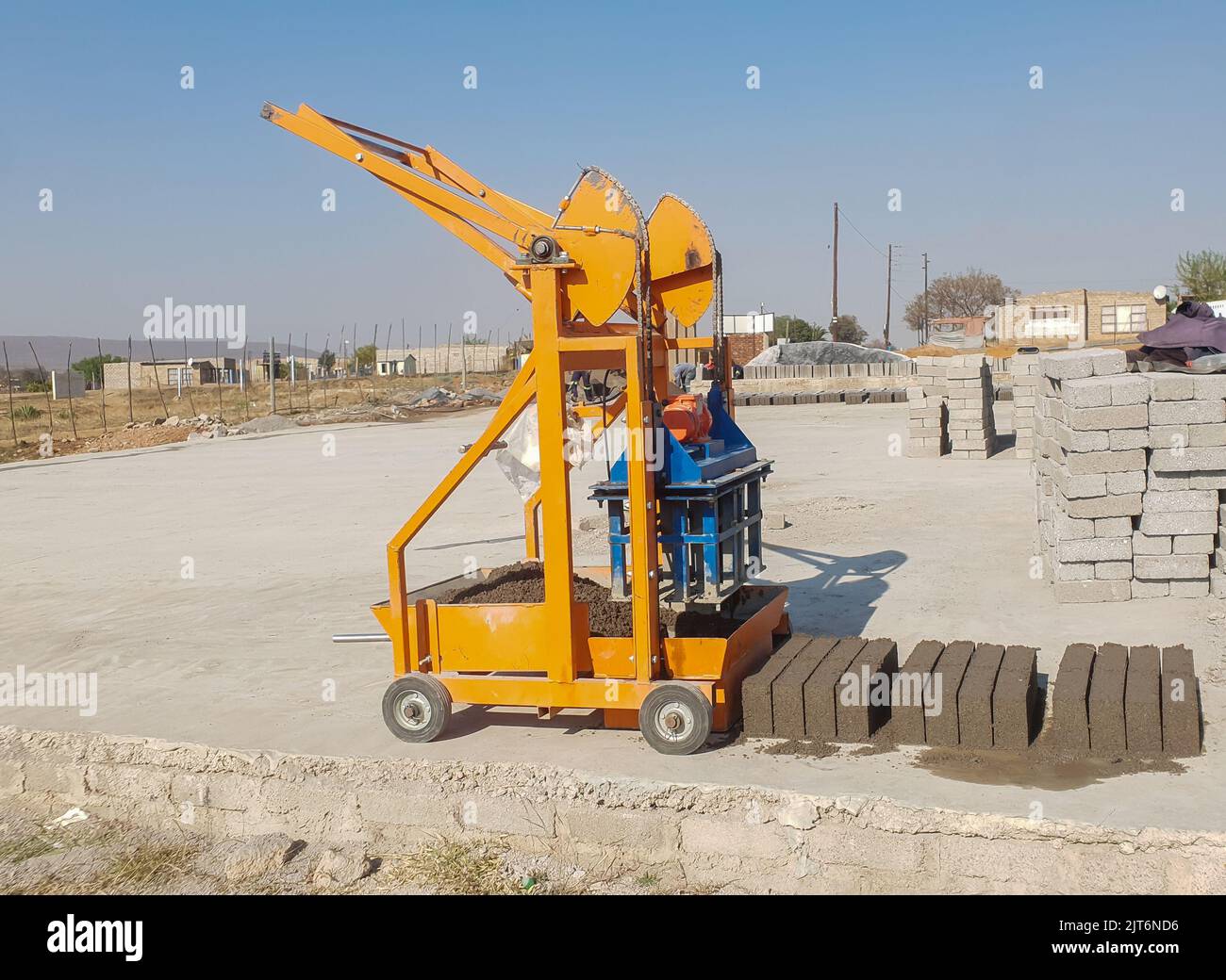 An orange metallic mobile brick making machine with wheels outside with fresh wet bricks next to the machine on a concrete floor. maxi brick mold with Stock Photo