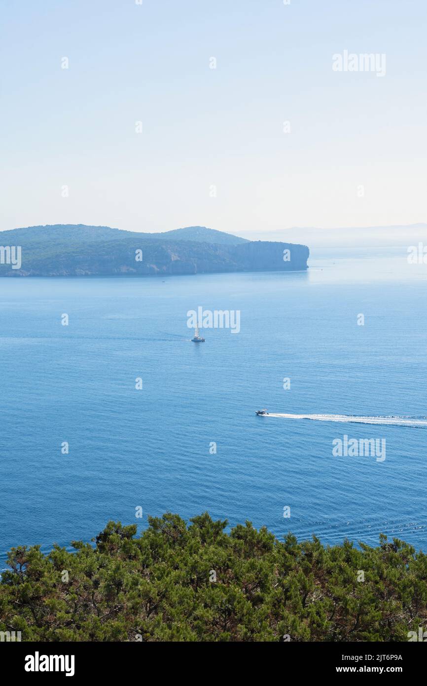 Vertical view from a hill of a cape in Italy with a sailboat in the distance and mountains in the background. Sunny midday with a turquoise Mediterran Stock Photo