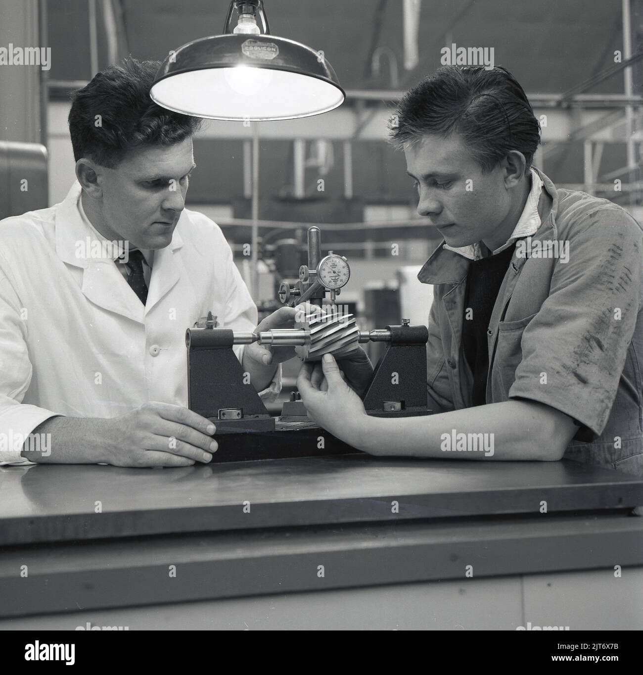 1950s, historical, aviation, engineering apprentice, sitting with his tutor, using a small piece of testing equipment with pressure gauge, Short Bros, Belfast, Northern Ireland, UK. An overhead industrial light made by Coolicon can be seen, first released in Britain in 1933 and a classic of British design. Stock Photo