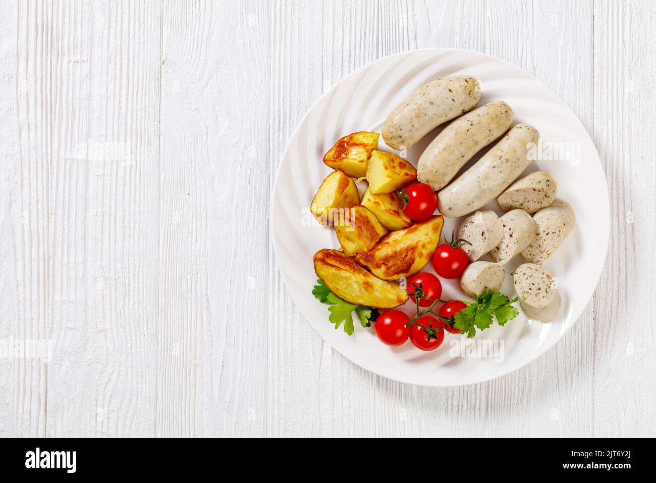 Weisswurst, bavarian white sausage of minced veal, pork back bacon, spices and parsley on white plate with roast potatoes, fresh tomatoes, horizontal Stock Photo