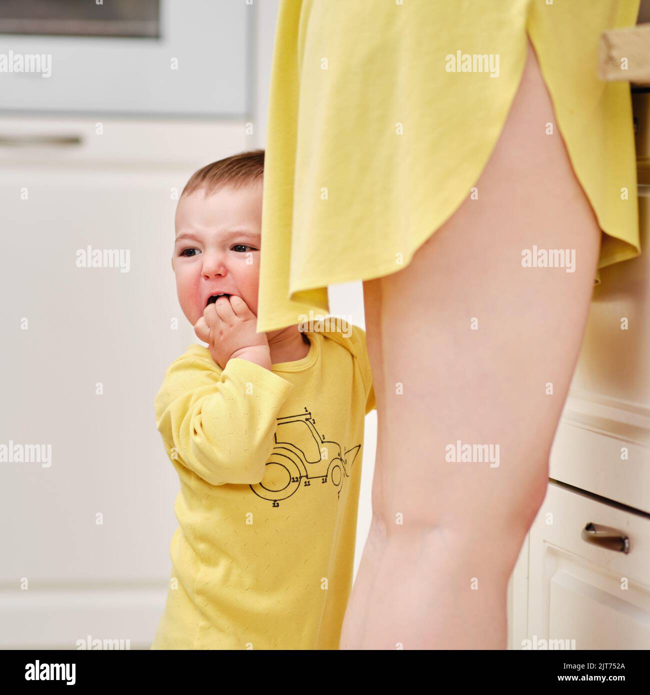 Crying toddler baby boy stands holding his mother legs in the kitchen